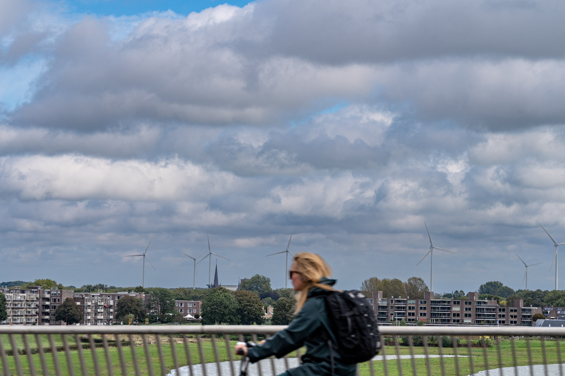 Een persoon met lang haar en een rugzak rijdt op een fiets over een brug. Op de achtergrond staan flatgebouwen, bomen, windturbines en een bewolkte lucht.
