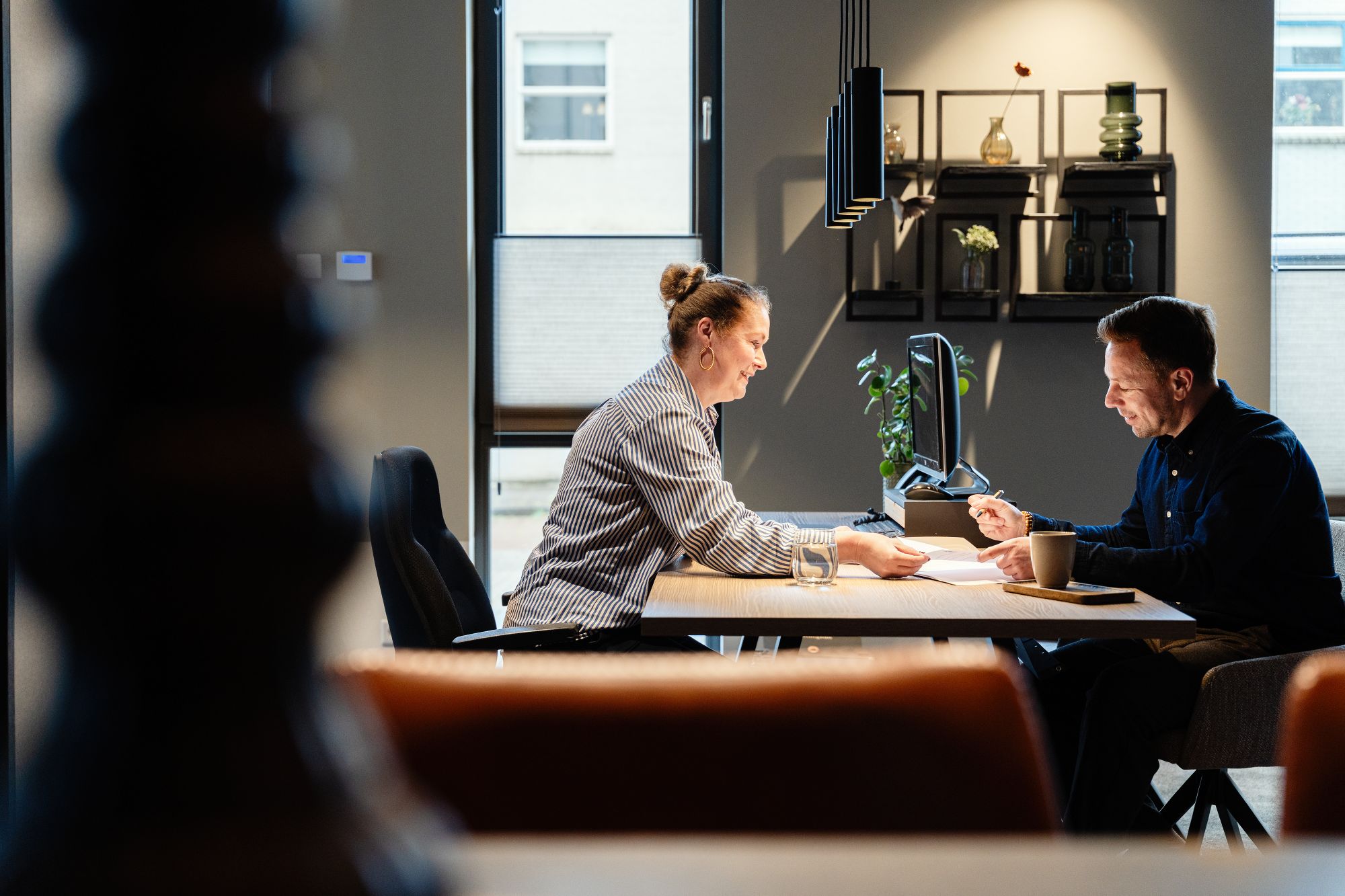 Twee mensen zitten tegenover elkaar aan een bureau in een modern kantoor en bespreken documenten. Zonlicht stroomt door het raam en creëert een warme, geconcentreerde sfeer. Planken en planten sieren de achtergrond.
