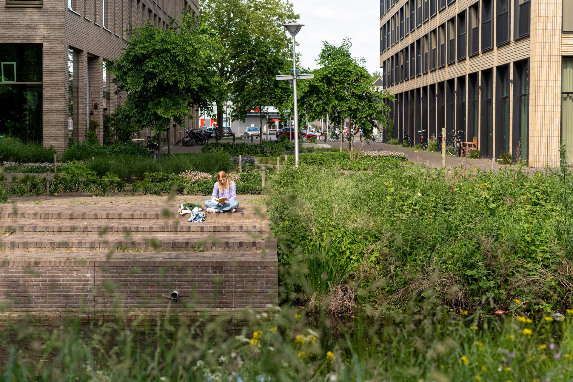 Een persoon zit alleen op een stenen trapje tussen het groen tussen twee moderne gebouwen, een boek te lezen met spullen in de buurt. Bomen en bloemen omringen het gebied en creëren een vredig stedelijk tafereel.