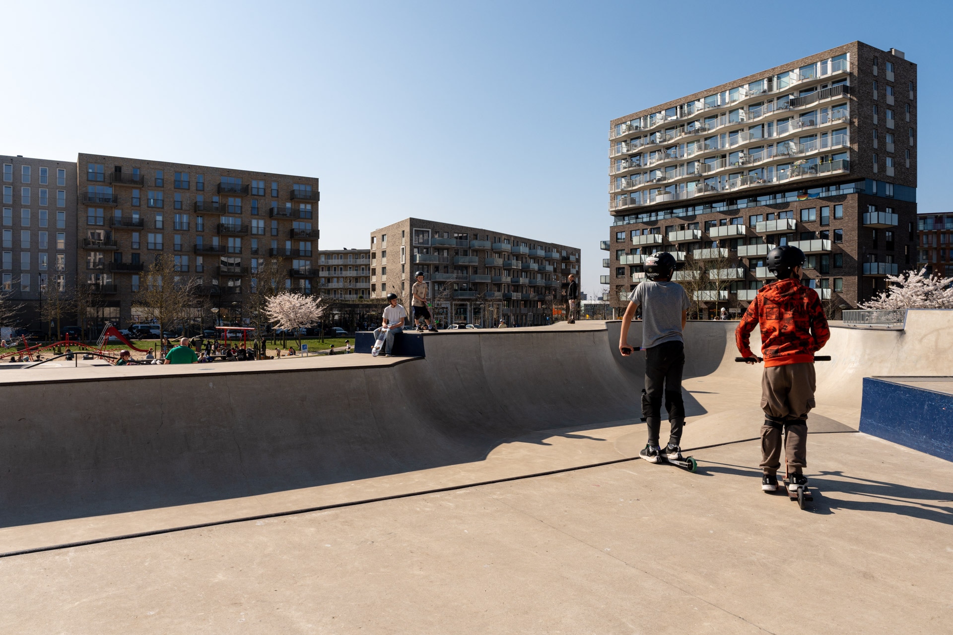 Twee kinderen met helmen op steppen in een skatepark in een stedelijke omgeving, omringd door moderne flatgebouwen en mensen die genieten van een zonnige dag.