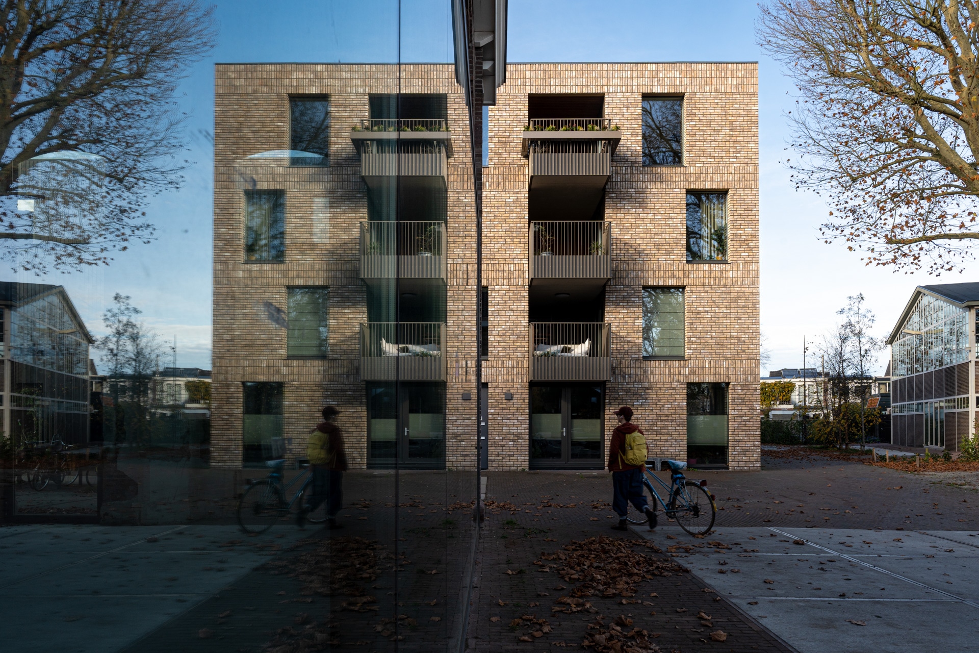 Een persoon met een fiets loopt langs een modern bakstenen flatgebouw, dat duidelijk weerspiegeld wordt in een groot glazen raam aan de linkerkant. Bladerloze bomen en afgevallen bladeren omzomen het geplaveide voetpad.