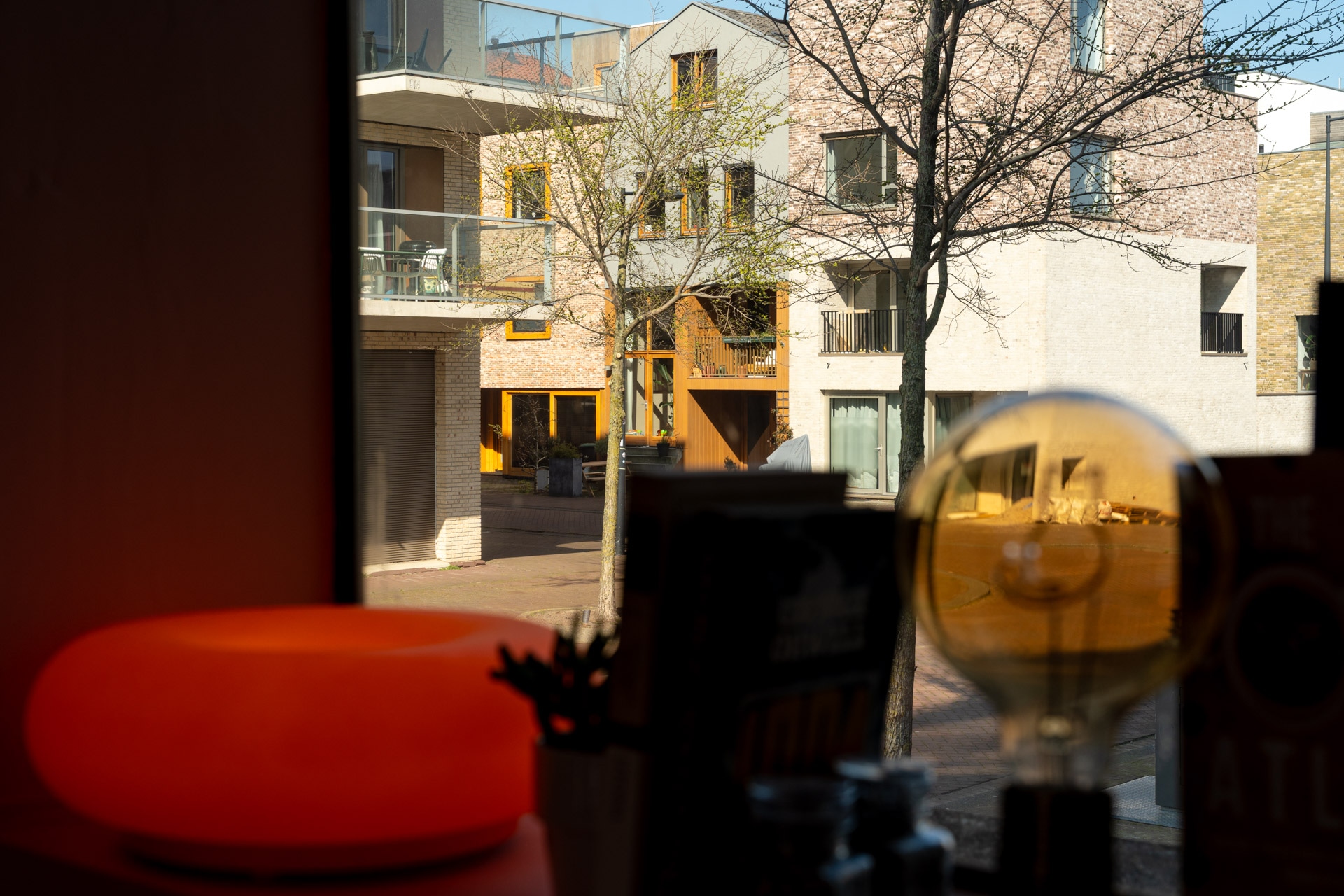 Een zonovergoten uitzicht door een raam toont moderne flatgebouwen, bladerloze bomen en een rustige straat. Binnen zijn een gloeiende oranje lamp, een gloeilamp en boeken zichtbaar op een vensterbank op de voorgrond.