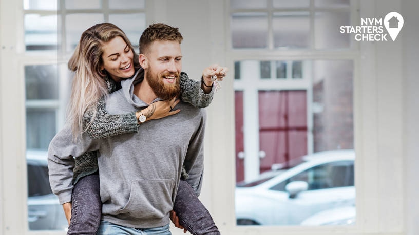 Een glimlachende vrouw rijdt binnenshuis op de rug van een bebaarde man. Beiden zijn casual gekleed en lijken gelukkig. Op de achtergrond zijn grote ramen en een geparkeerde auto zichtbaar. Het NVM Starters Check logo staat in de rechterbovenhoek.