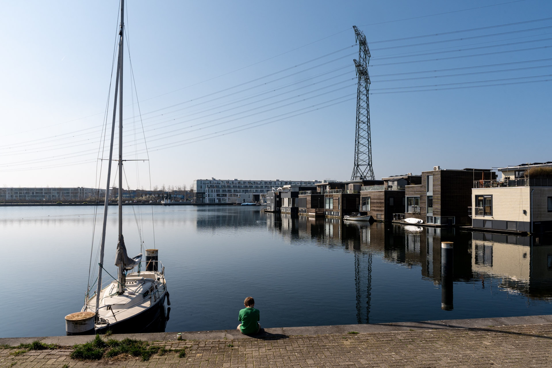 Een kind in een groen shirt zit aan een kalme waterkant bij een aangemeerde zeilboot. Moderne woonboten dobberen op het water en op de achtergrond verrijst een hoogspanningsmast met elektriciteitsdraden onder een strakblauwe hemel.