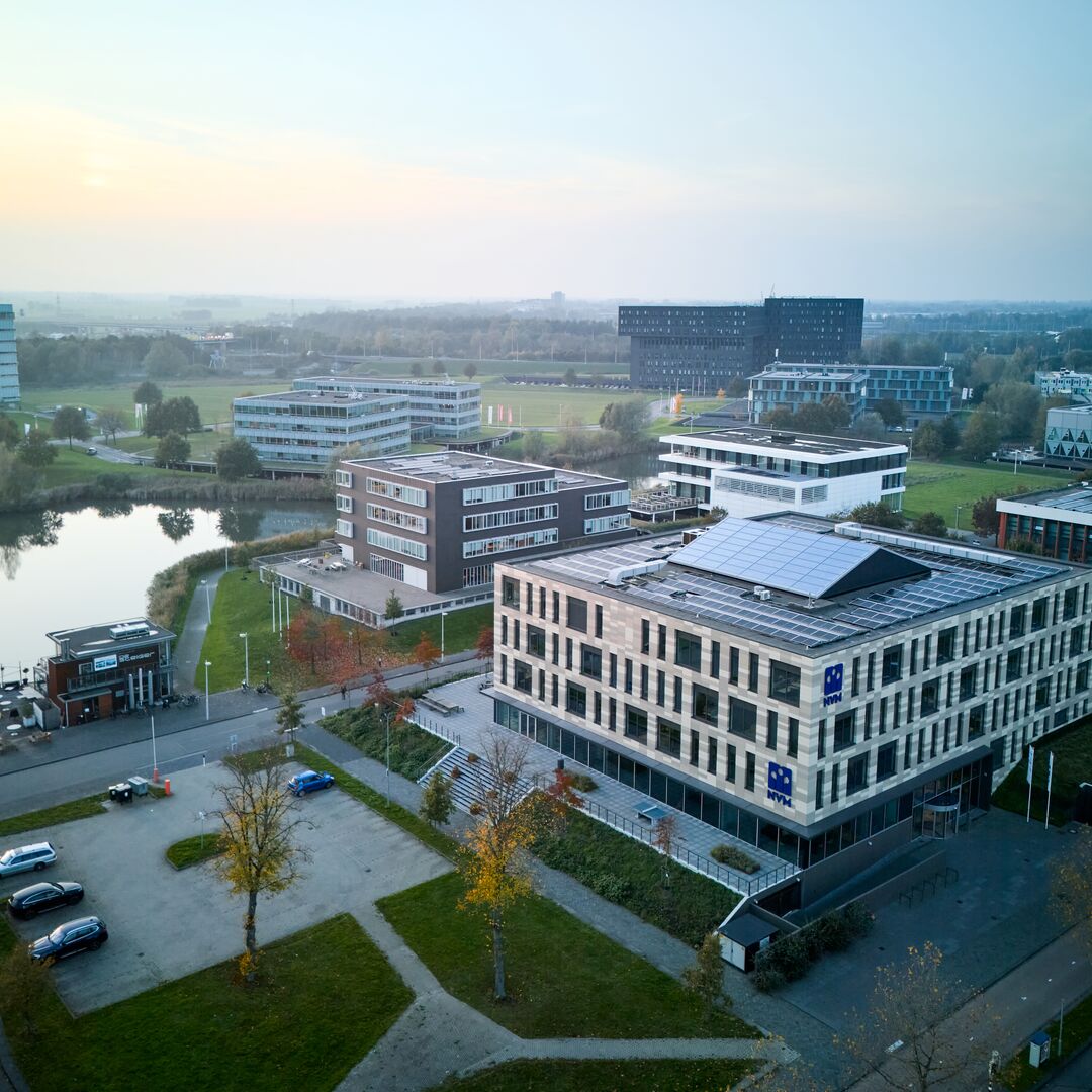 Luchtfoto van een moderne campus met verschillende rechthoekige gebouwen, zonnepanelen op daken, een kleine parkeerplaats en een vijver omgeven door groene gazons en bomen onder een wazige hemel.