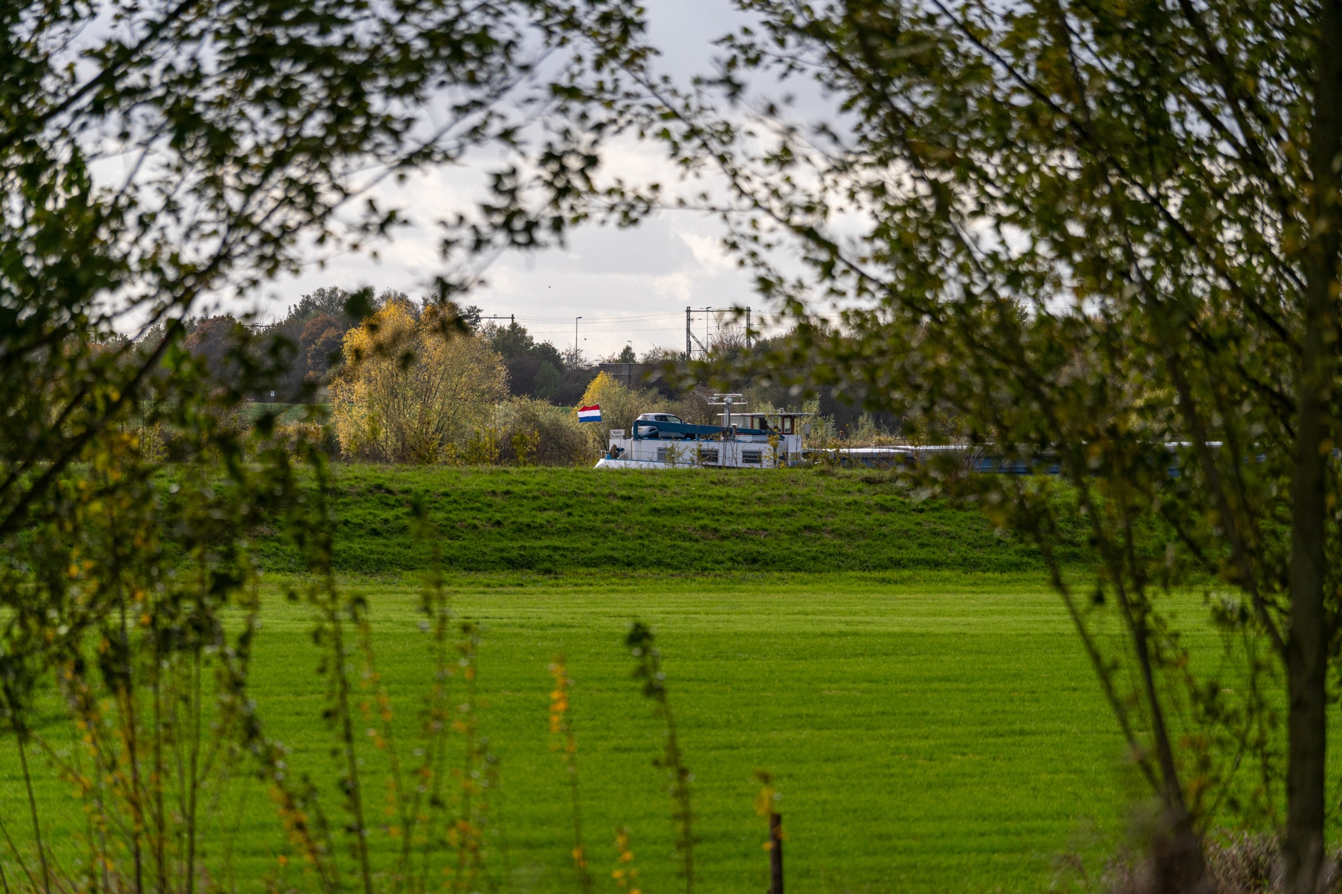 Een vrachtschip met een Nederlandse vlag vaart achter een met gras begroeide dijk, omlijst door bomen en groene velden onder een bewolkte hemel.