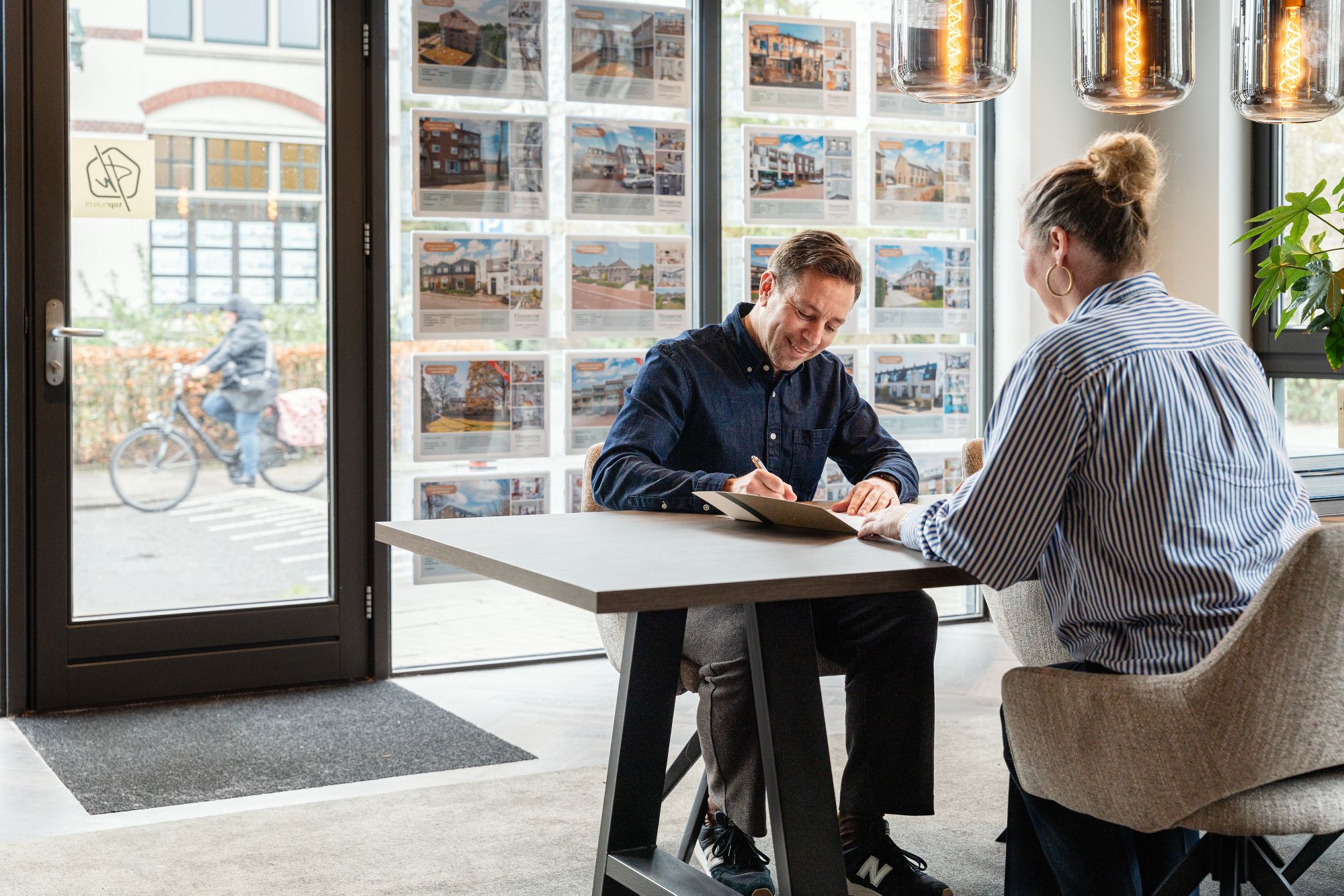 Een man en een vrouw zitten aan een tafel in een modern kantoor met grote ramen. De man ondertekent papieren terwijl de vrouw toekijkt. Buiten rijdt iemand op een fiets langs het raam met onroerend goed advertenties.