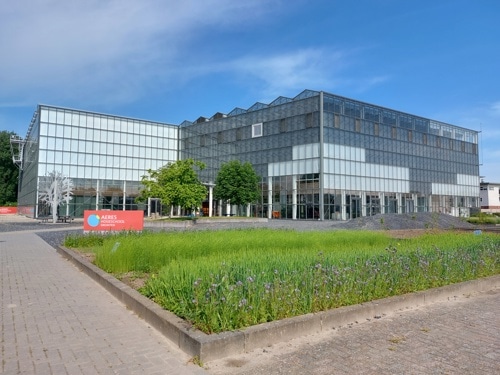 Een modern glazen gebouw met een rastervormige gevel, omgeven door een geplaveid voetpad en groene beplanting. Een rood bord met de tekst Aeres staat bij de ingang. De lucht is helder en blauw.