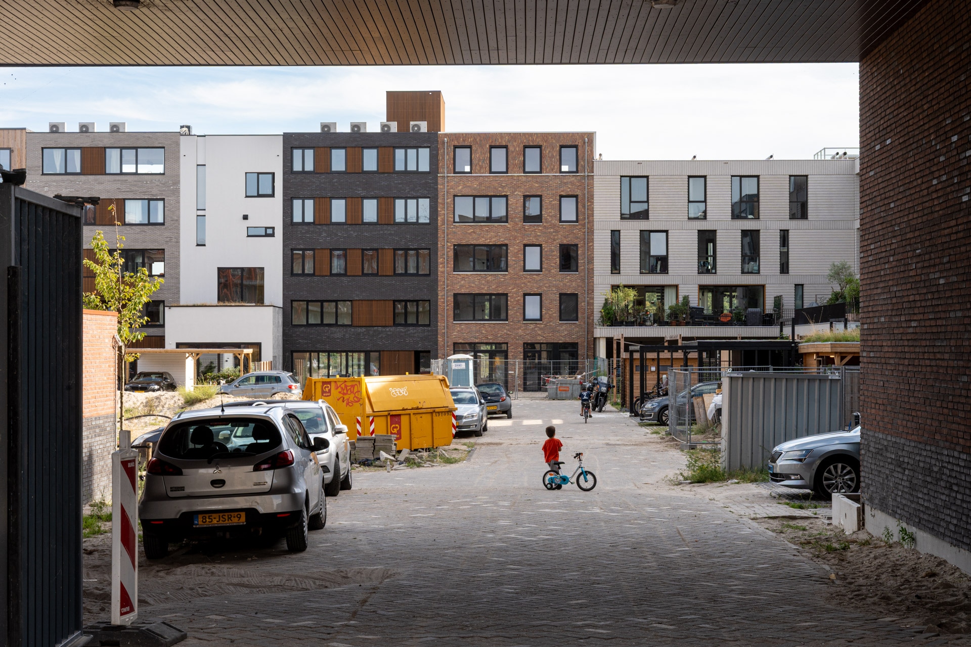 Een jong kind op een blauwe fiets rijdt in de richting van een woonwijk met moderne flatgebouwen, geparkeerde auto's, een gele afvalcontainer en bouwmaterialen zichtbaar op een gedeeltelijk zanderige weg.
