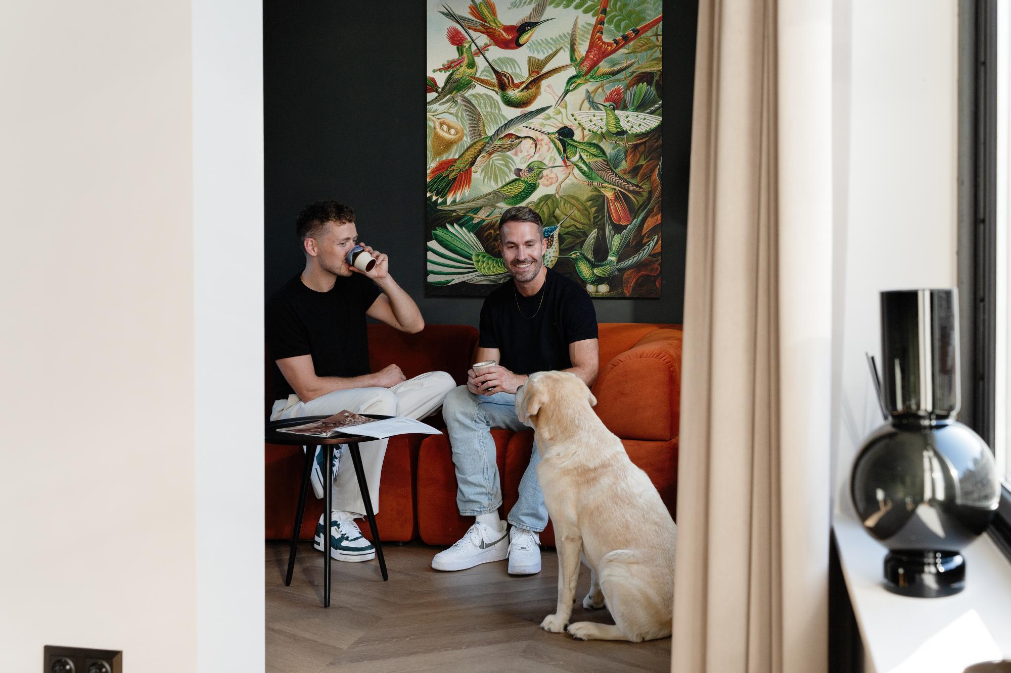 Twee mannen zitten op een oranje bank, glimlachen en houden koffiekopjes vast, met voor hen een gele hond. Achter hen hangt een kleurrijk botanisch schilderij op een donkere muur. Vlakbij staan een ronde tafel en een tijdschrift.