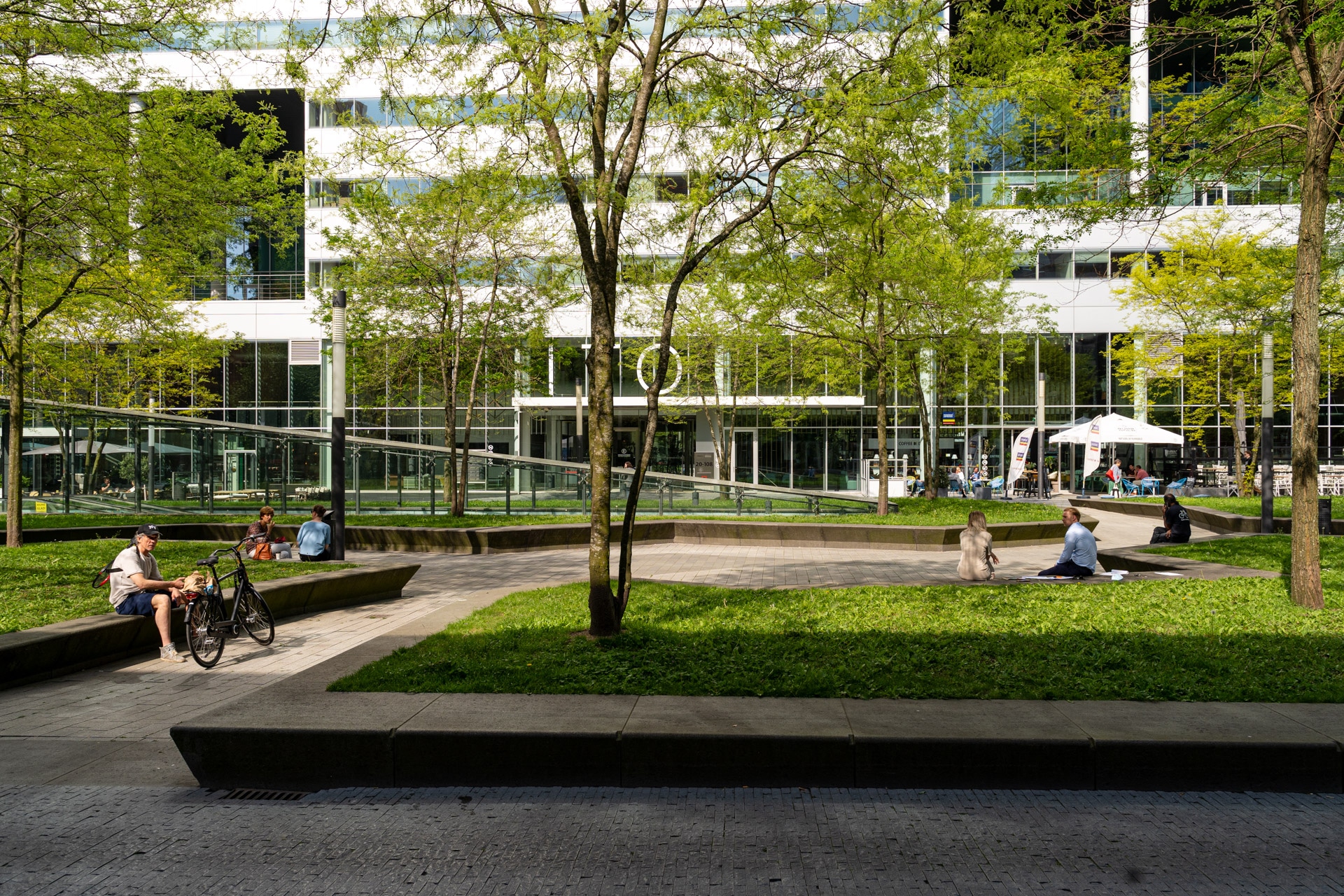 Mensen zitten en ontspannen op bankjes en gras in een moderne stedelijke binnenplaats met bomen en glazen gebouwen op de achtergrond op een zonnige dag.