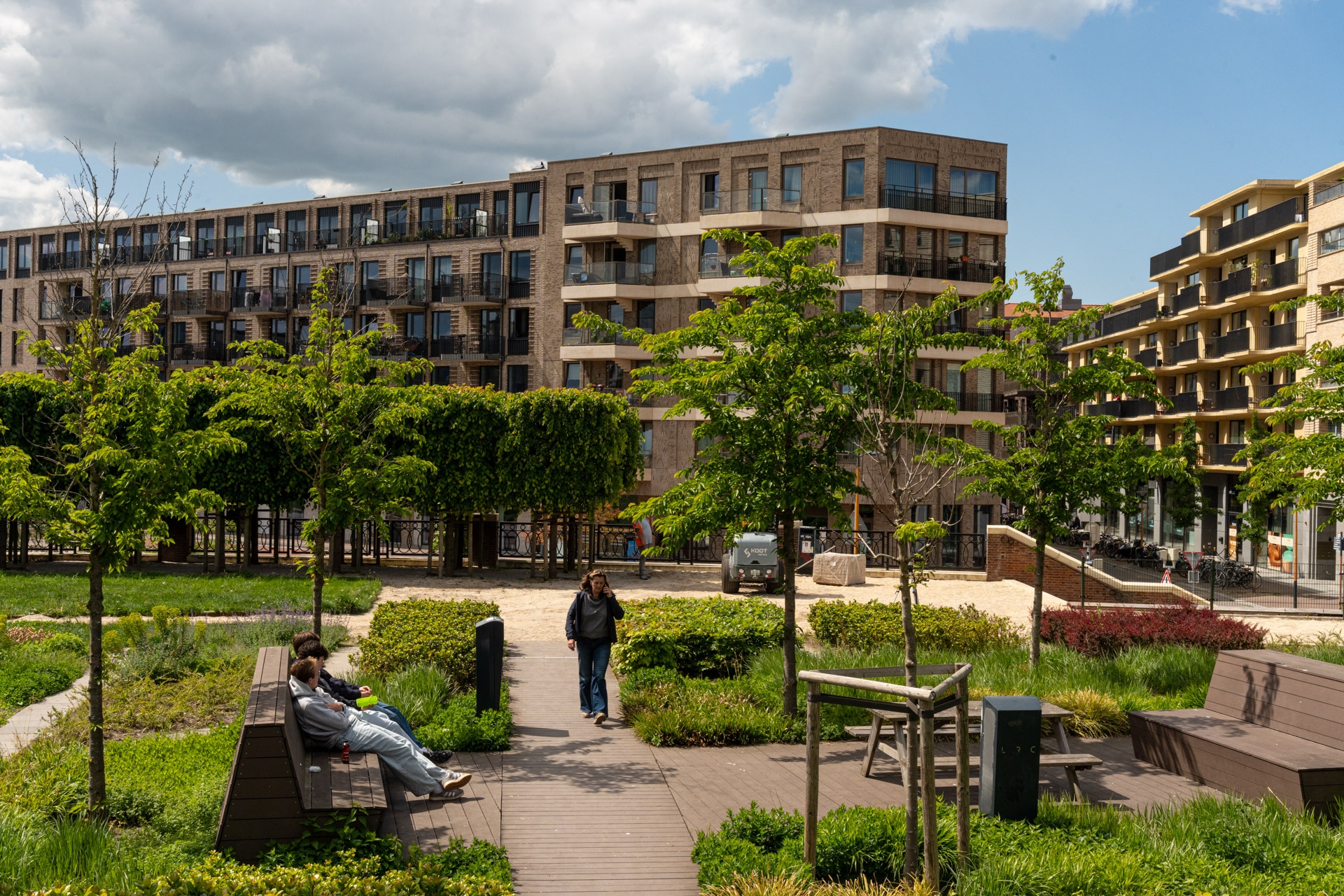 Mensen ontspannen in een modern stadspark met bankjes, groen en jonge bomen. Flatgebouwen met balkons omringen het park onder een gedeeltelijk bewolkte hemel.