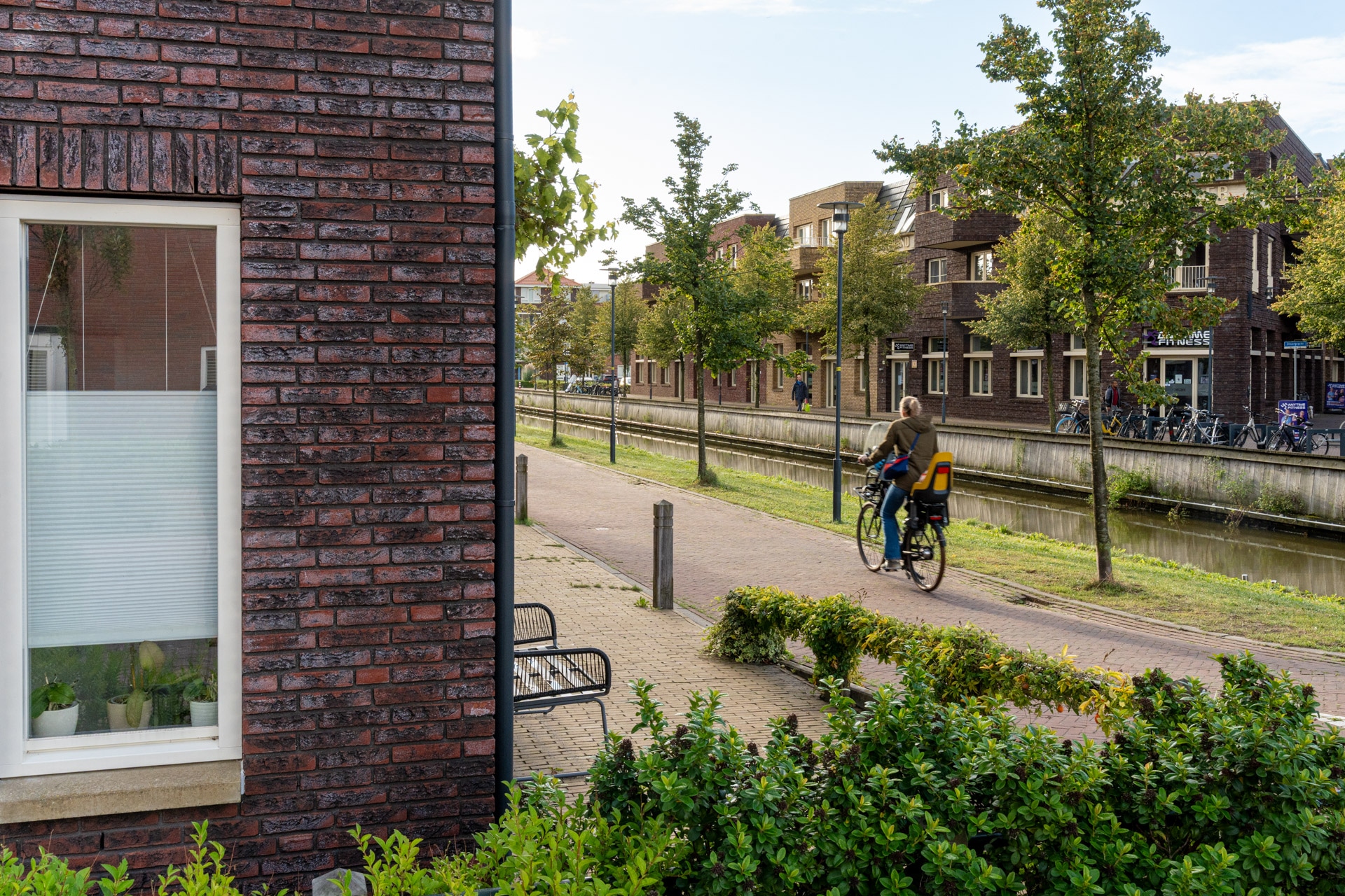 Een persoon fietst op een heldere dag over een pad langs de gracht in een woonwijk met bakstenen gebouwen, bomen en groen.