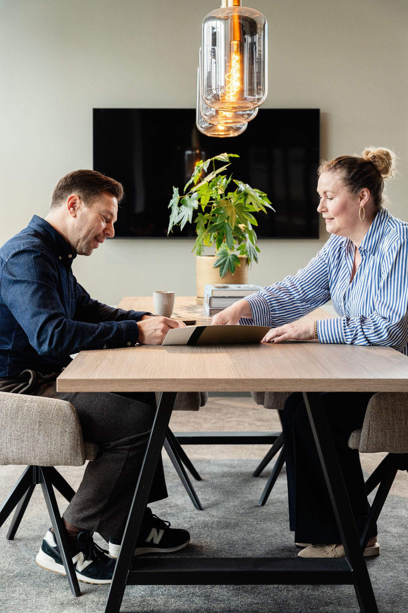 Een man en een vrouw zitten tegenover elkaar aan een houten tafel en kijken naar een tablet. Een potplant en een koffiemok staan op tafel, met een moderne lamp en tv op de achtergrond.