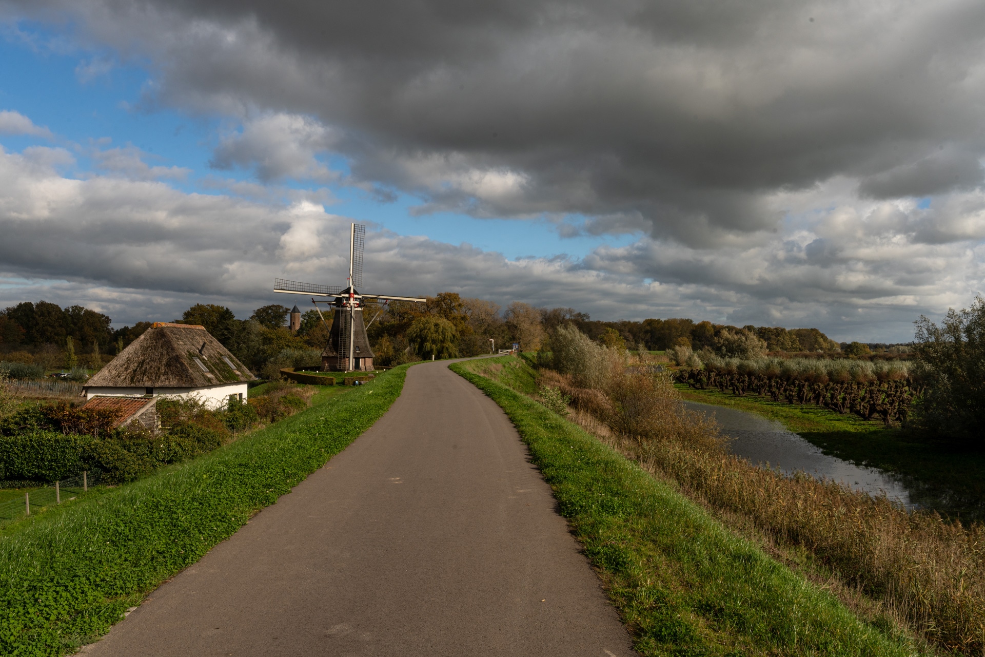 Een verharde weg kronkelt door groene velden naar een traditionele Nederlandse windmolen en een huis met rieten dak, met dramatische wolken en flarden zonlicht boven je hoofd.