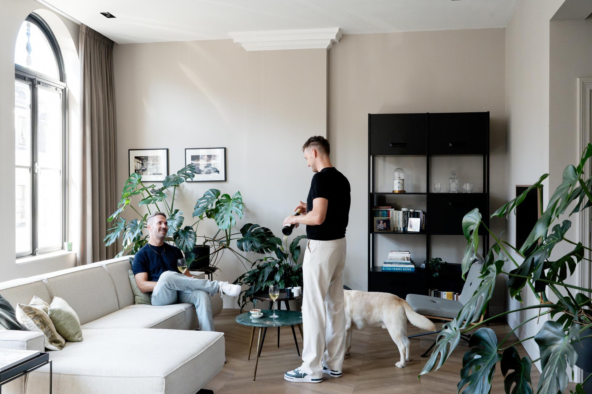 Twee mannen en een hond bevinden zich in een lichte woonkamer met grote planten, een beige bank, een ronde glazen salontafel en een zwarte kast. Eén man zit op de bank terwijl de ander staat, met de hond naast zich.