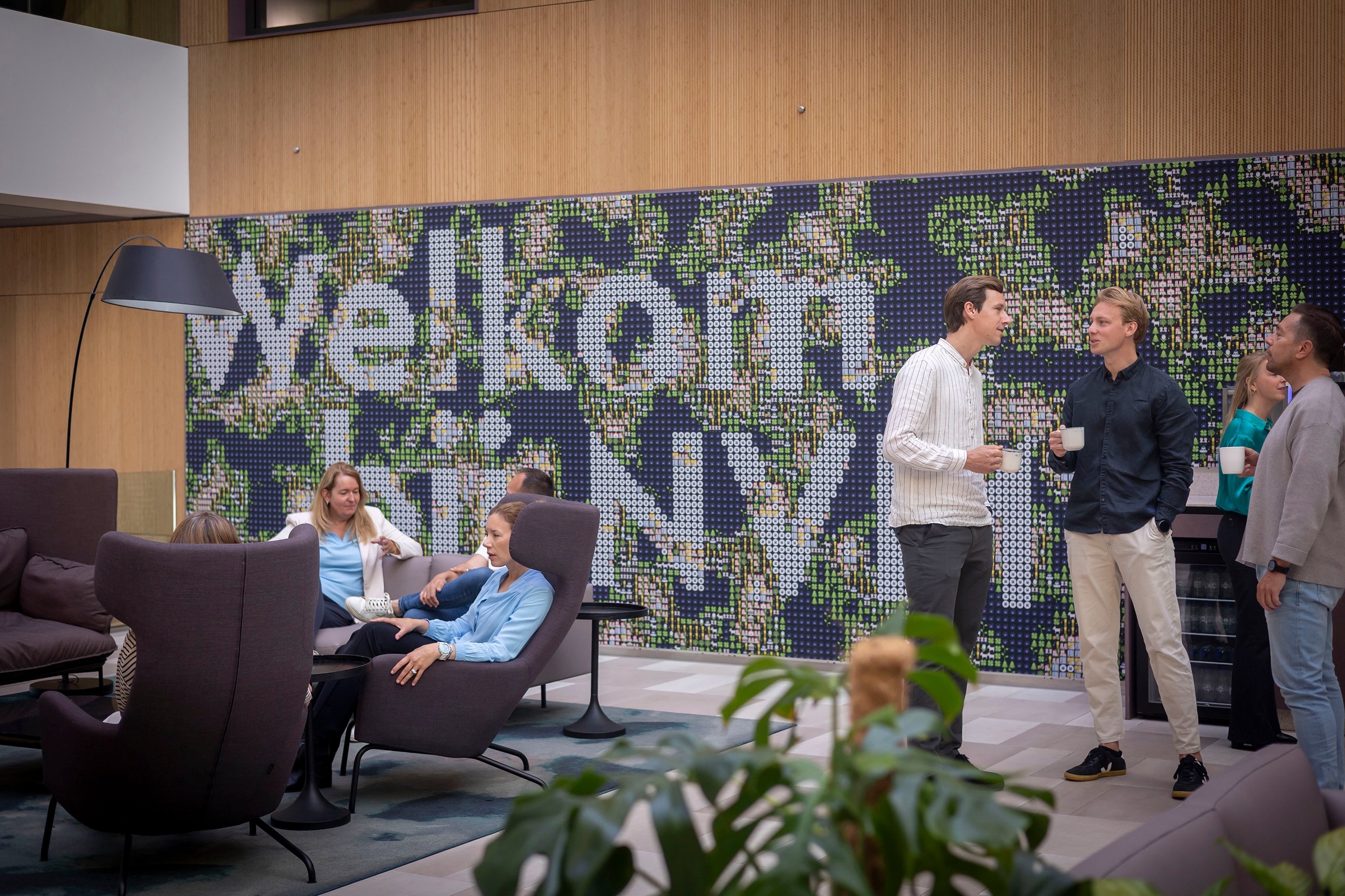Een groep mensen maakt kennis met elkaar in een moderne loungeruimte bij een groene muur met het woord Welkom in grote letters. Sommigen zitten en anderen staan, houden kopjes vast en kletsen in een ontspannen sfeer.