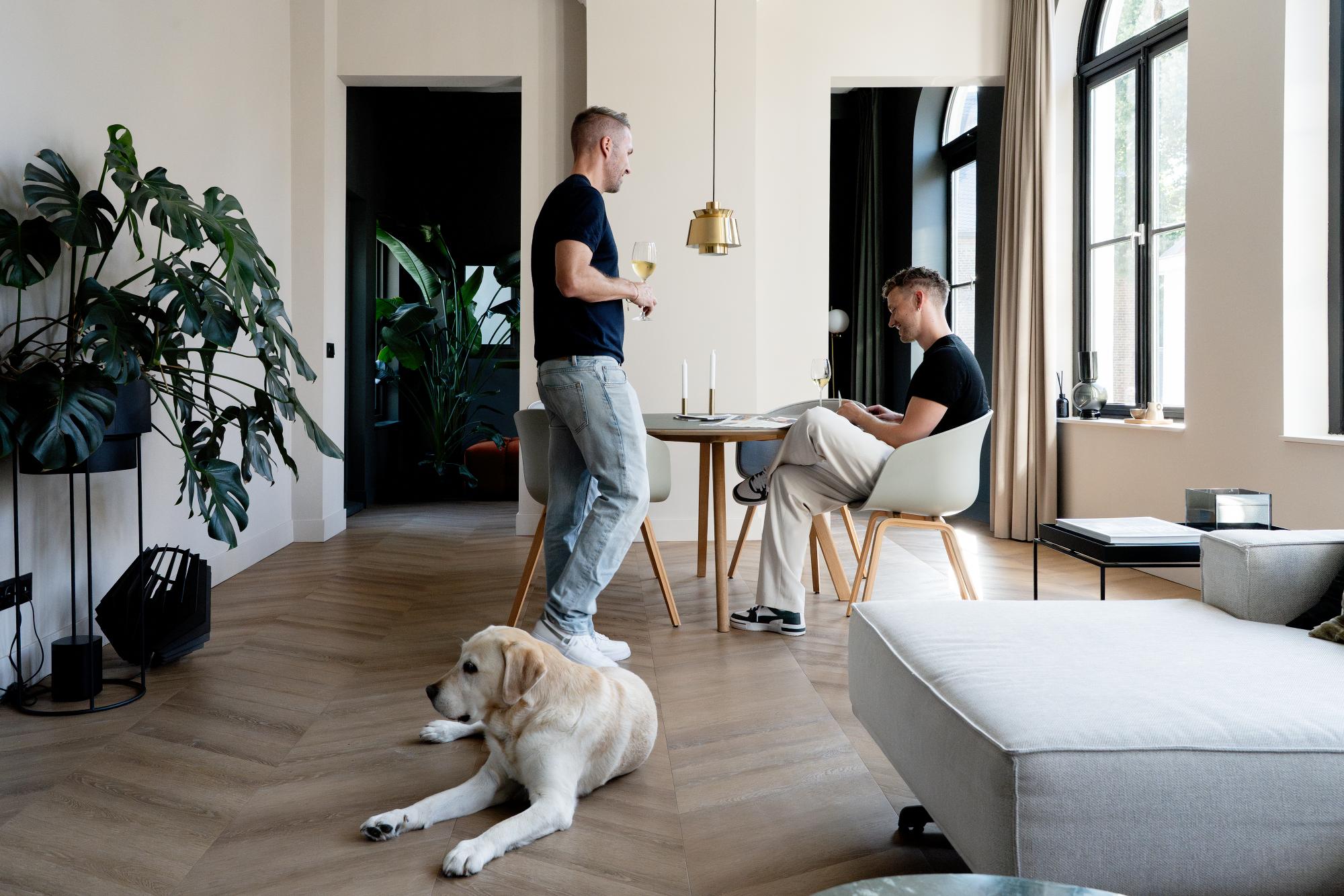 Een man staat met een drankje in zijn hand en praat met een andere man die met een laptop aan een eettafel zit. Een gele labrador ligt op de houten vloer in een moderne, lichte leefruimte met planten en grote ramen.