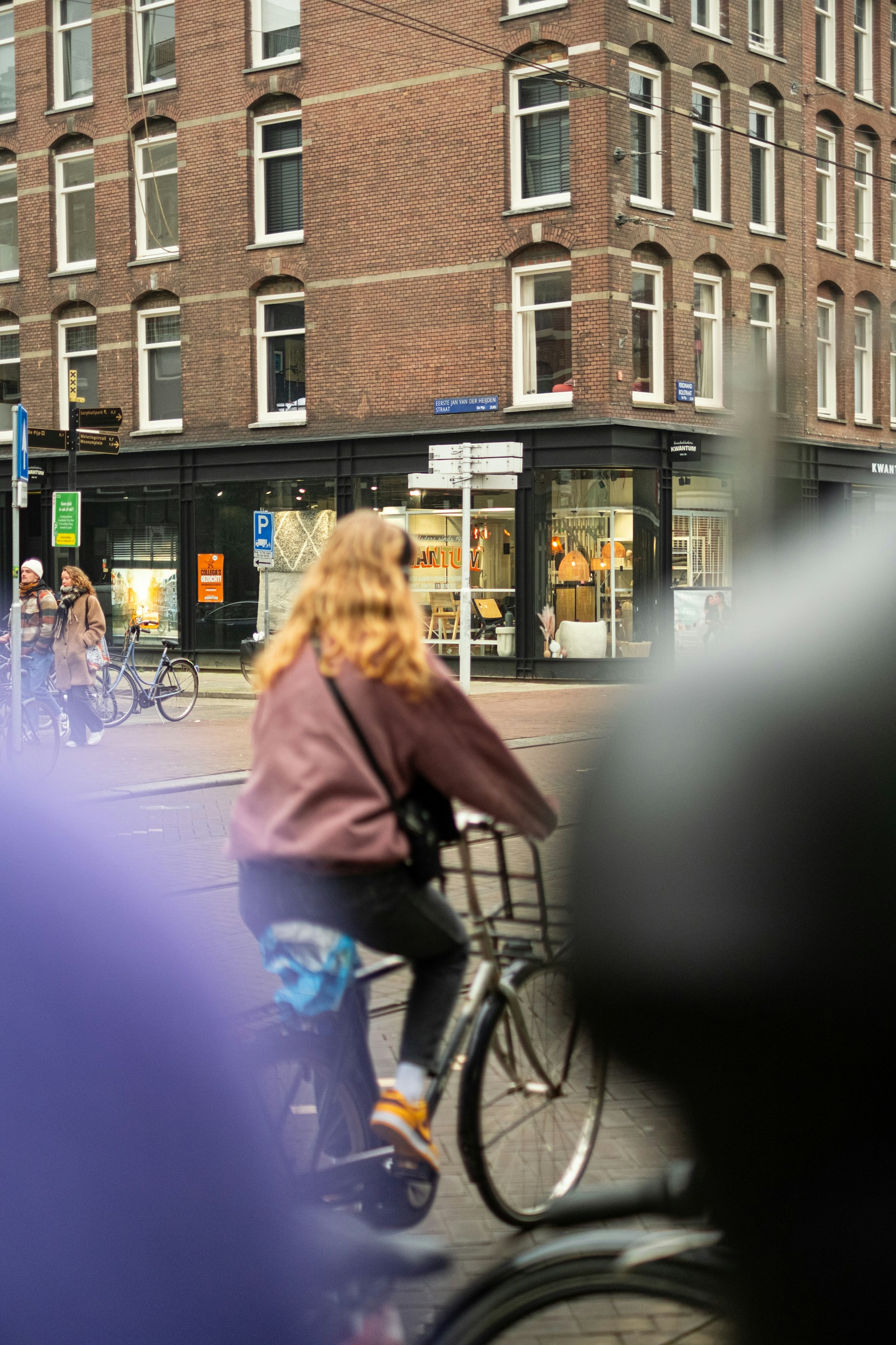 Een persoon met krullend haar fietst op een stadsstraat voor een bakstenen gebouw, maar het beeld is sterk vervormd met digitale haperingen en kleurstrepen.