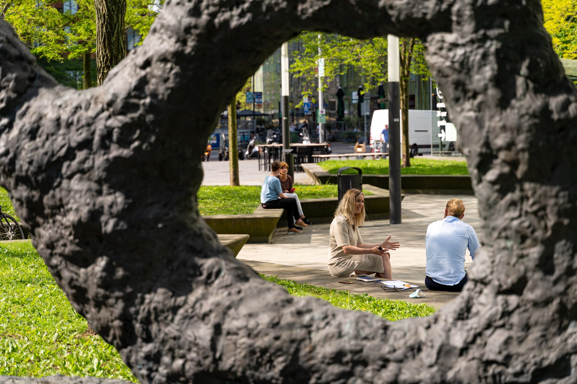 Drie mensen zitten op bankjes en op het gras in een zonnig park, omlijst door een cirkelvormige opening in een sculptuur met donkere textuur op de voorgrond. Bomen en groen omringen het tafereel.