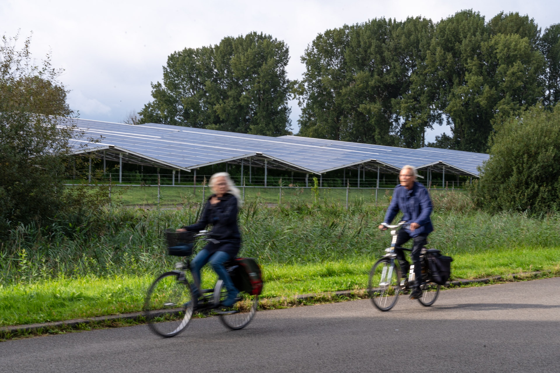 Twee mensen fietsen op een pad naast een veld met zonnepanelen, met hoge groene bomen en een bewolkte lucht op de achtergrond.