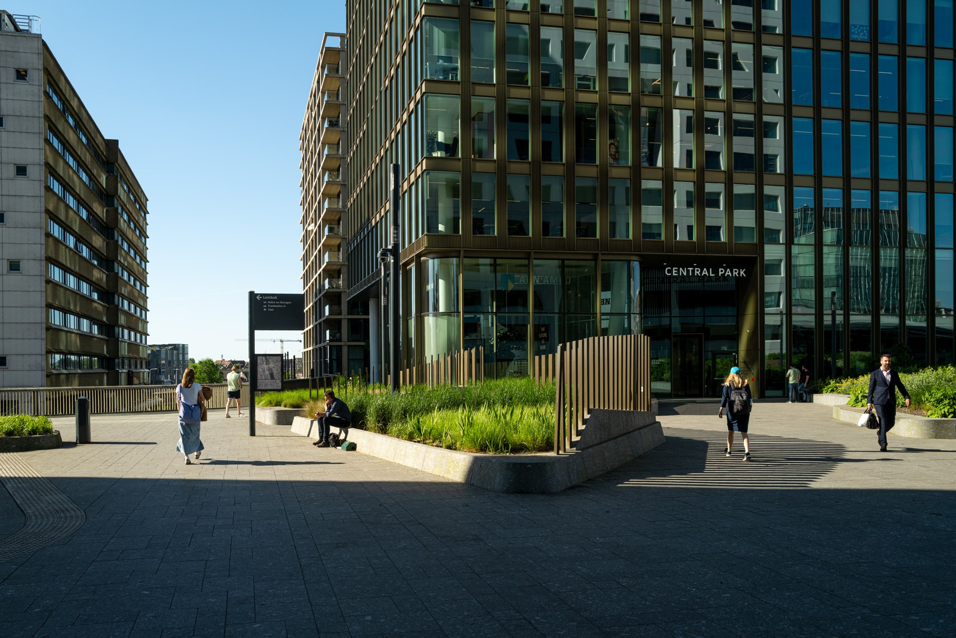 Mensen lopen en zitten op een zonnige dag bij een modern glazen kantoorgebouw dat Central Park heet; groen omzoomt de ingang en schaduwen vallen over het trottoir.