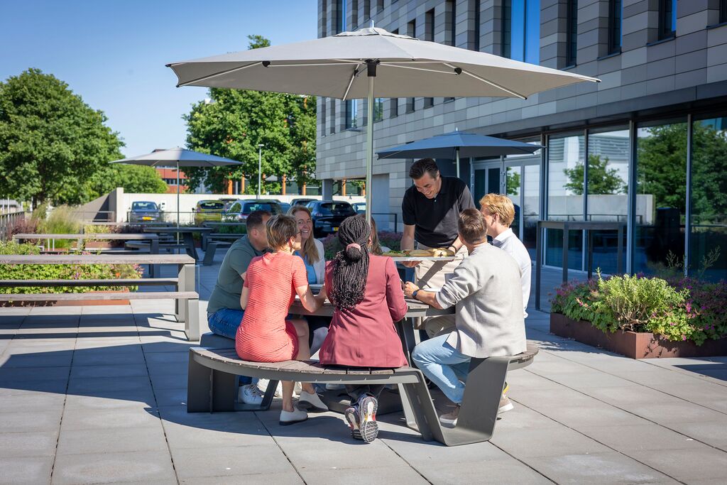 Een groep mensen zit rond een ronde tafel onder een grote paraplu, in gesprek buiten een modern gebouw op een zonnige dag.