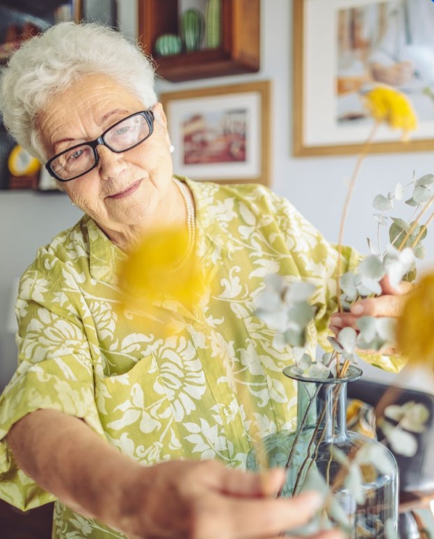 Een oudere vrouw met wit haar en een bril, draagt een groen bloemetjeshemd en schikt gele bloemen in een glazen vaas binnenshuis. Op de achtergrond hangen ingelijste foto's aan de muur.