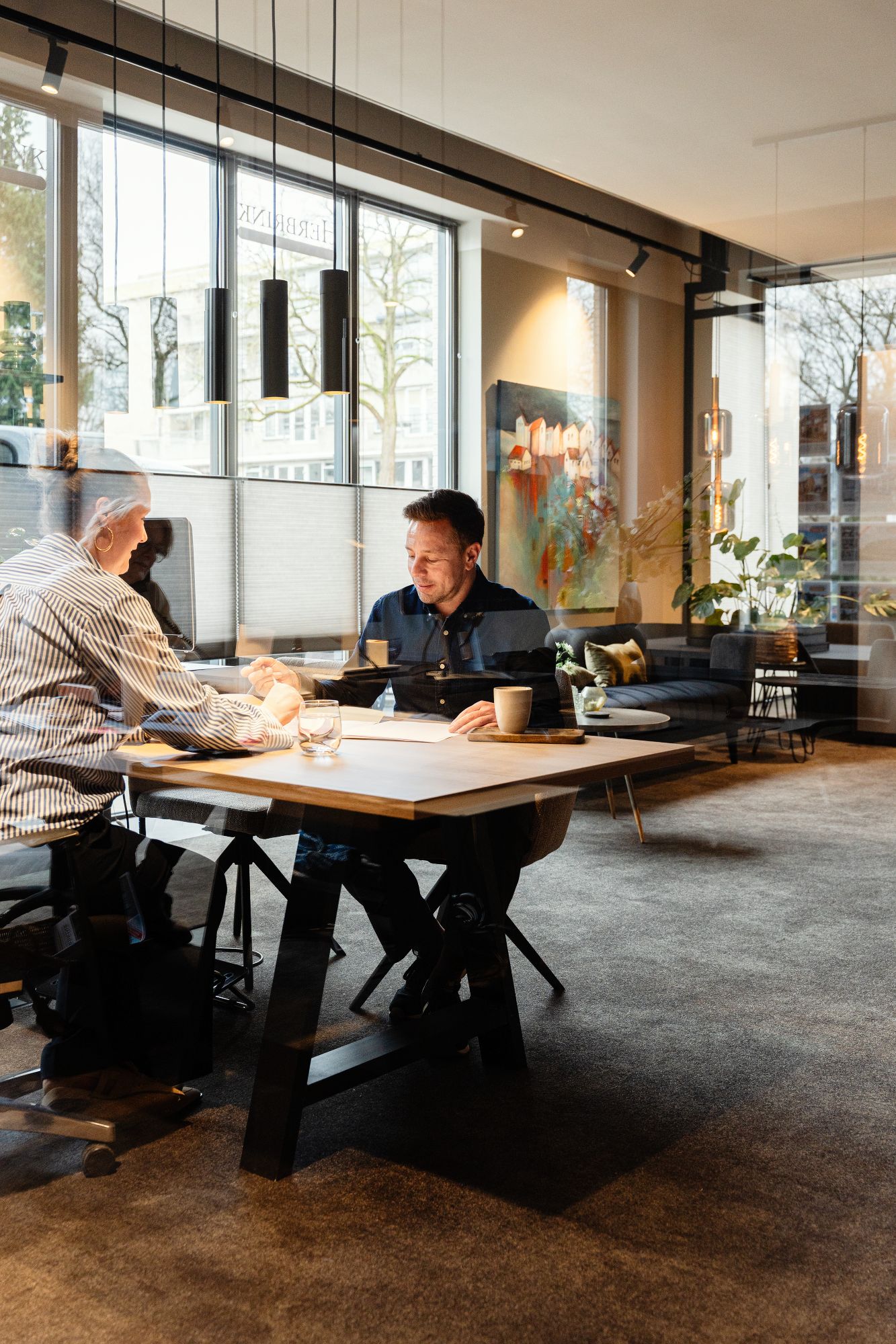 Twee mensen zitten aan een moderne kantoortafel met papieren en koffiekopjes en zijn in gesprek. Grote ramen laten natuurlijk licht binnen en op de achtergrond is een eigentijdse inrichting met planten en kunstwerken te zien.