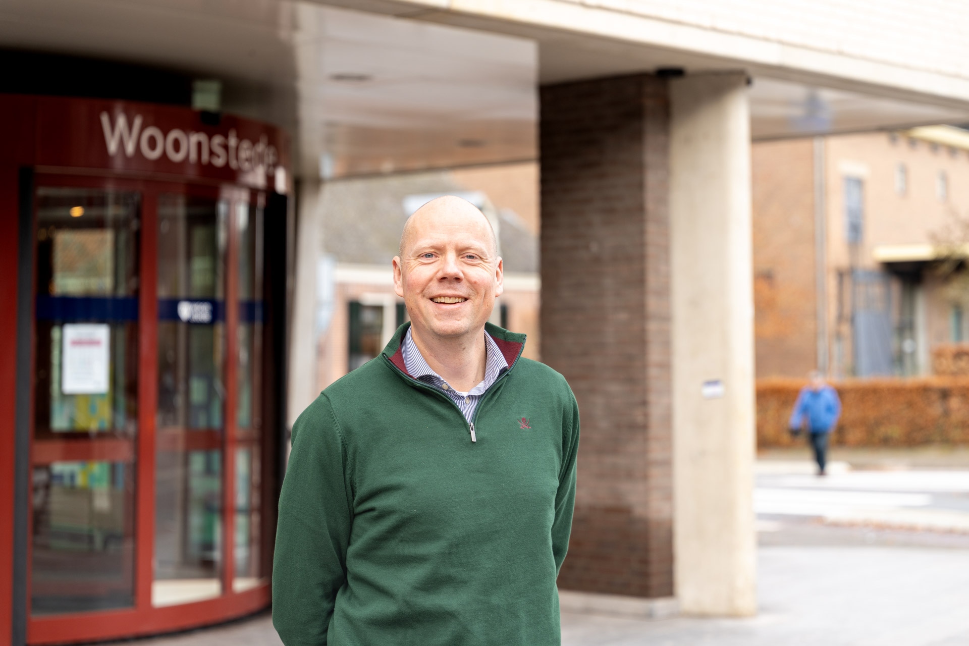 Een lachende man in een groene trui staat voor een gebouw met een glazen ingang met de tekst Woonstede. Op de achtergrond loopt een persoon in het blauw op het trottoir in de buurt van enkele bakstenen gebouwen en herfstbladeren.