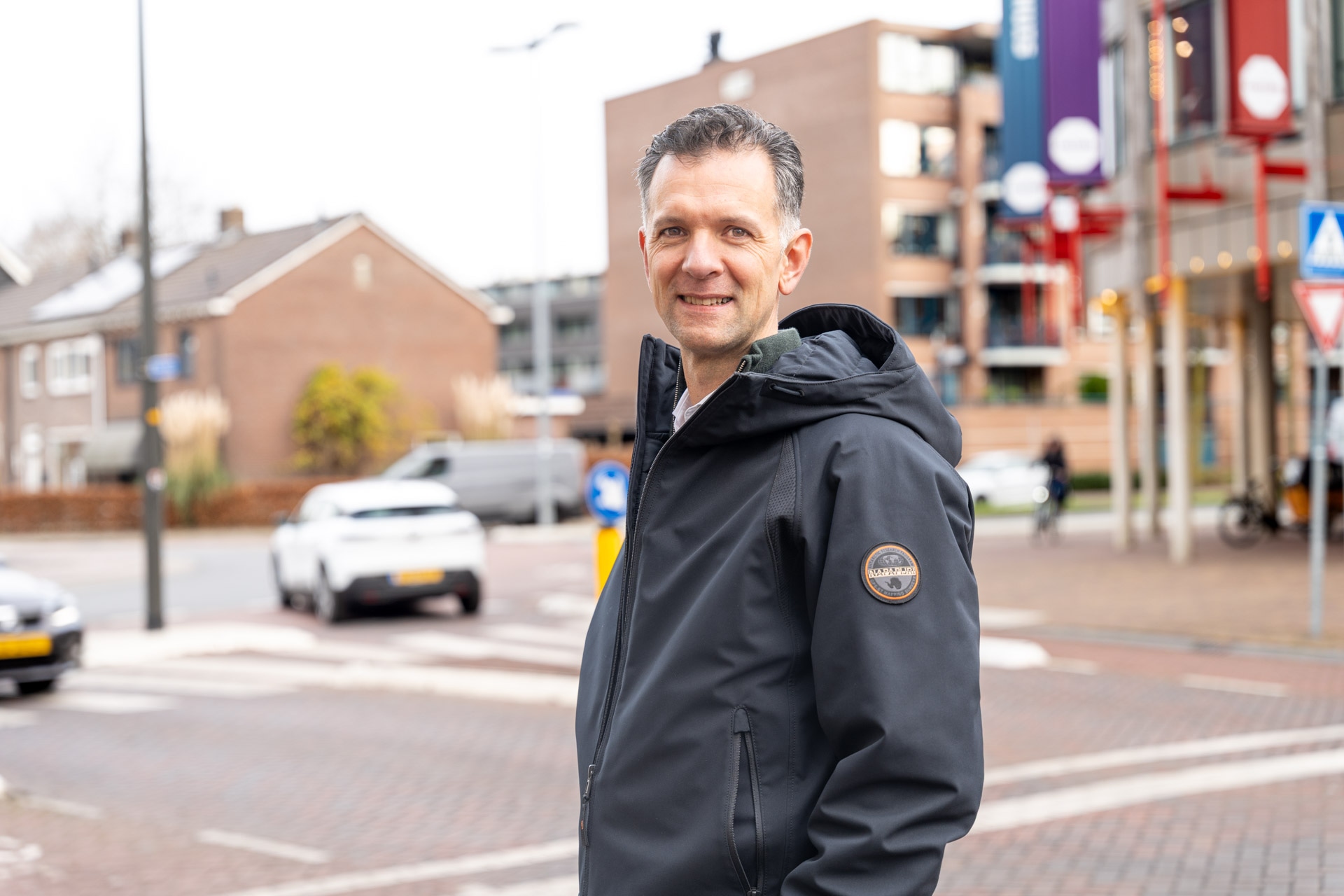 Een man met een donker jack staat op de hoek van een straat en lacht naar de camera. Gebouwen, auto's en een fiets zijn te zien op de achtergrond op een bewolkte dag.