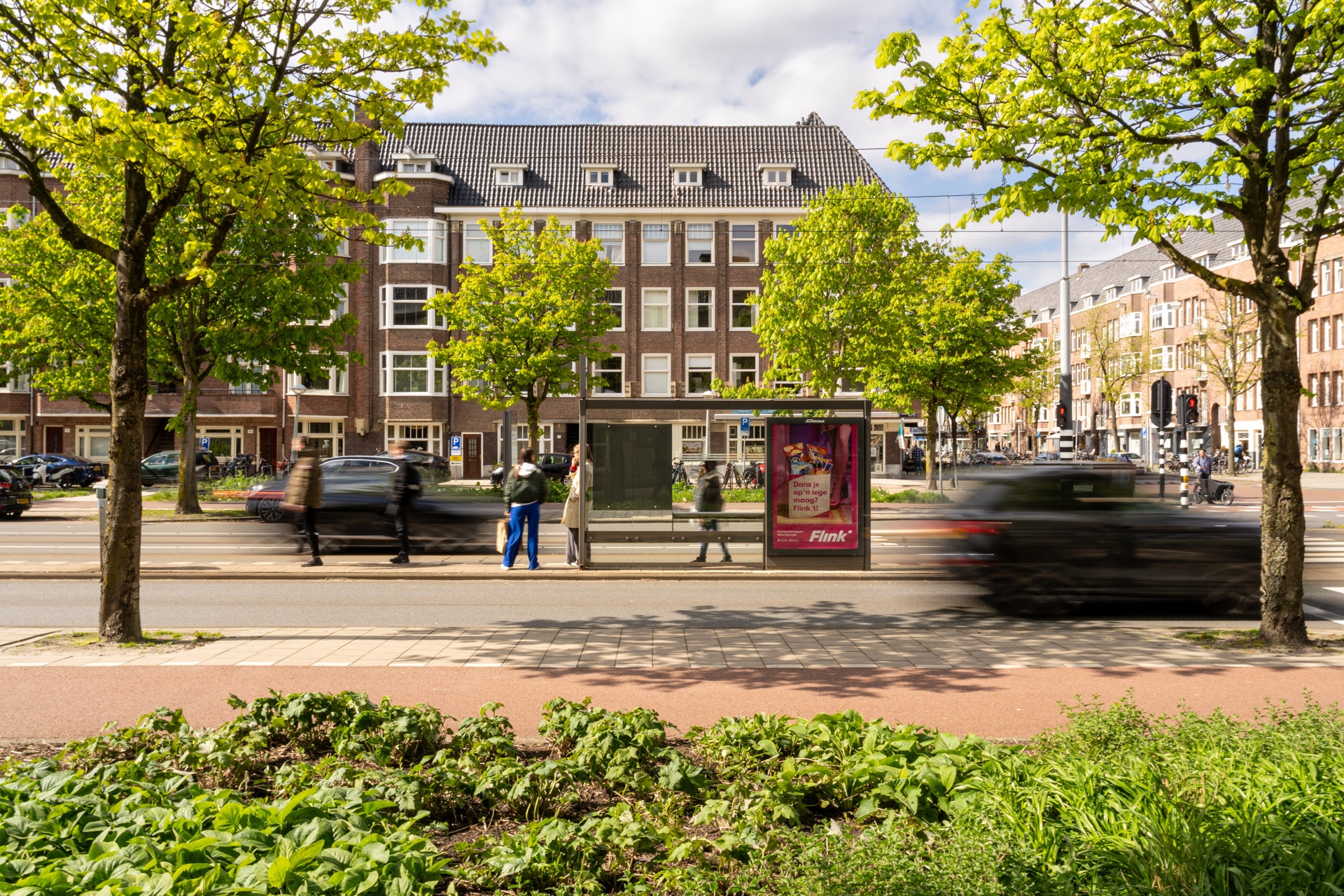 Een stadsstraat met wachtende mensen bij een bushalte, wazig voorbijrijdende auto's en bomen op het trottoir voor bakstenen flatgebouwen op een zonnige dag.