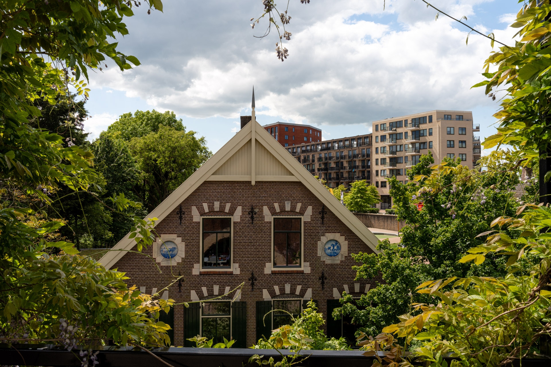 Een traditioneel bakstenen huis met decoratieve tegels wordt omringd door groene bomen, met op de achtergrond moderne appartementsgebouwen onder een gedeeltelijk bewolkte hemel.