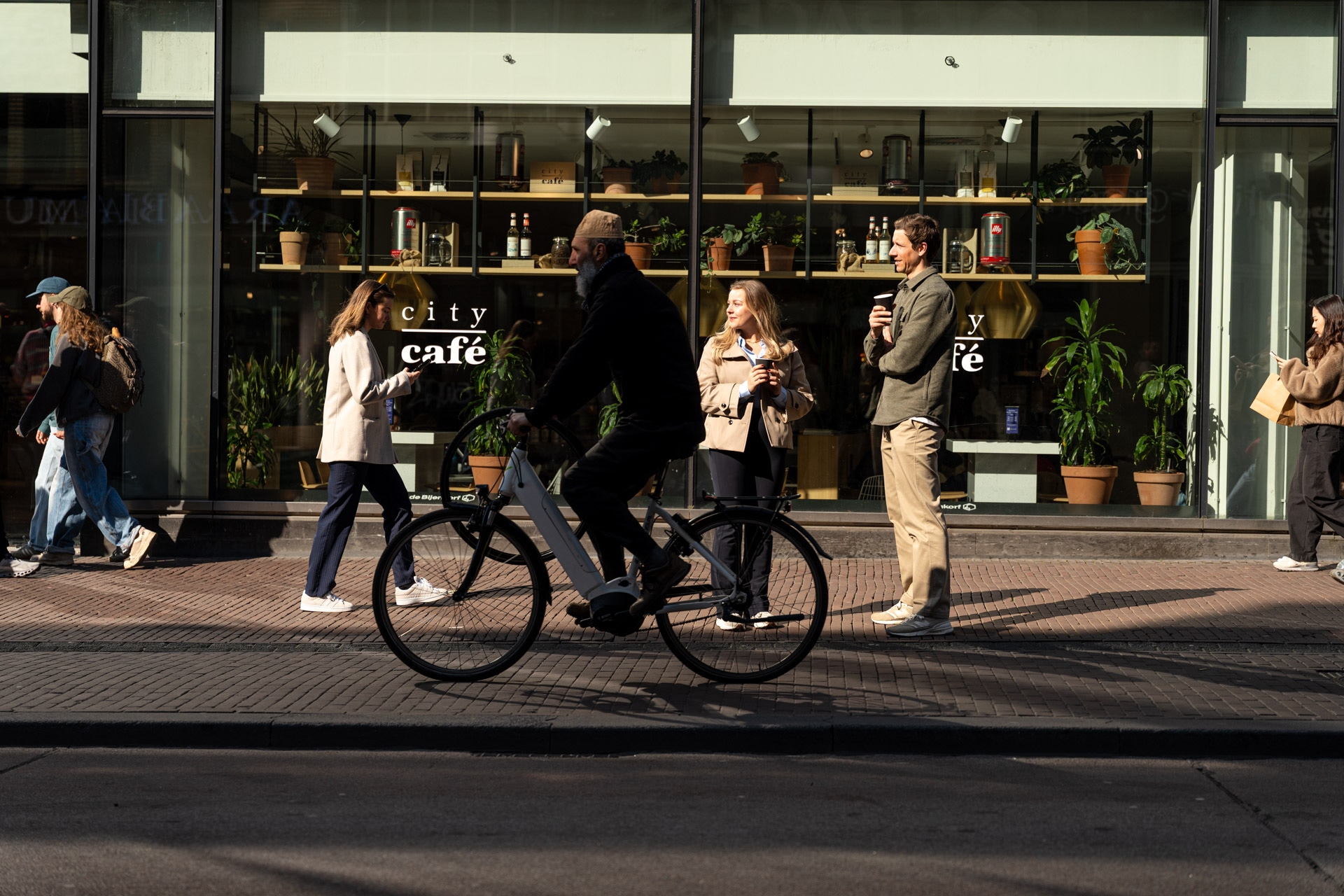Een persoon rijdt op een fiets langs verschillende lopende en staande mensen buiten een café met grote glazen ramen vol planten en planken, in een zonnige stadsstraat.