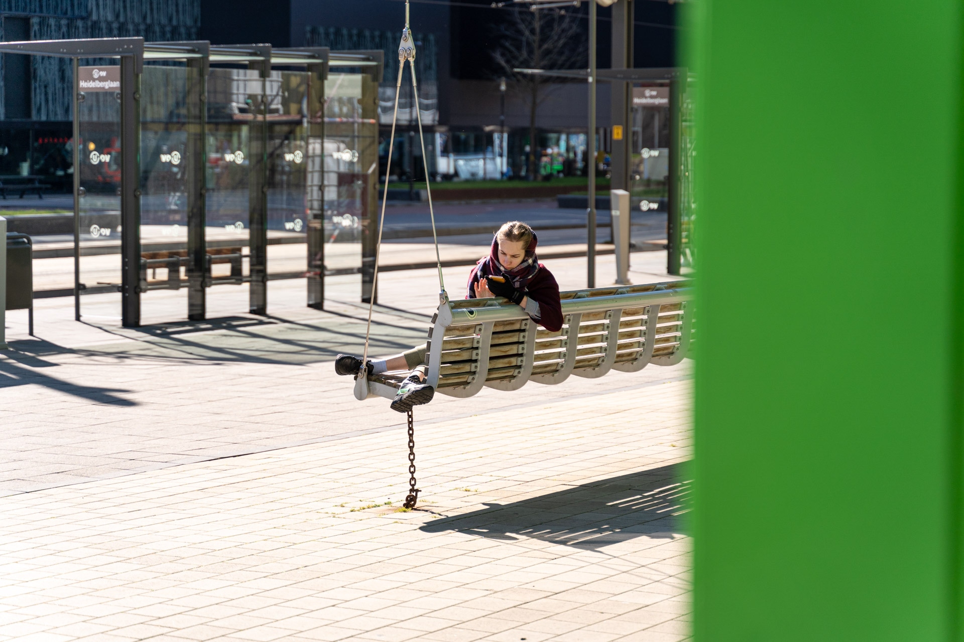 Een kind zit op een hangende schommelbank bij een moderne bushalte buiten een boek te lezen. Zonlicht werpt schaduwen op de betegelde grond. Rechts op de voorgrond is een grote groene structuur zichtbaar.