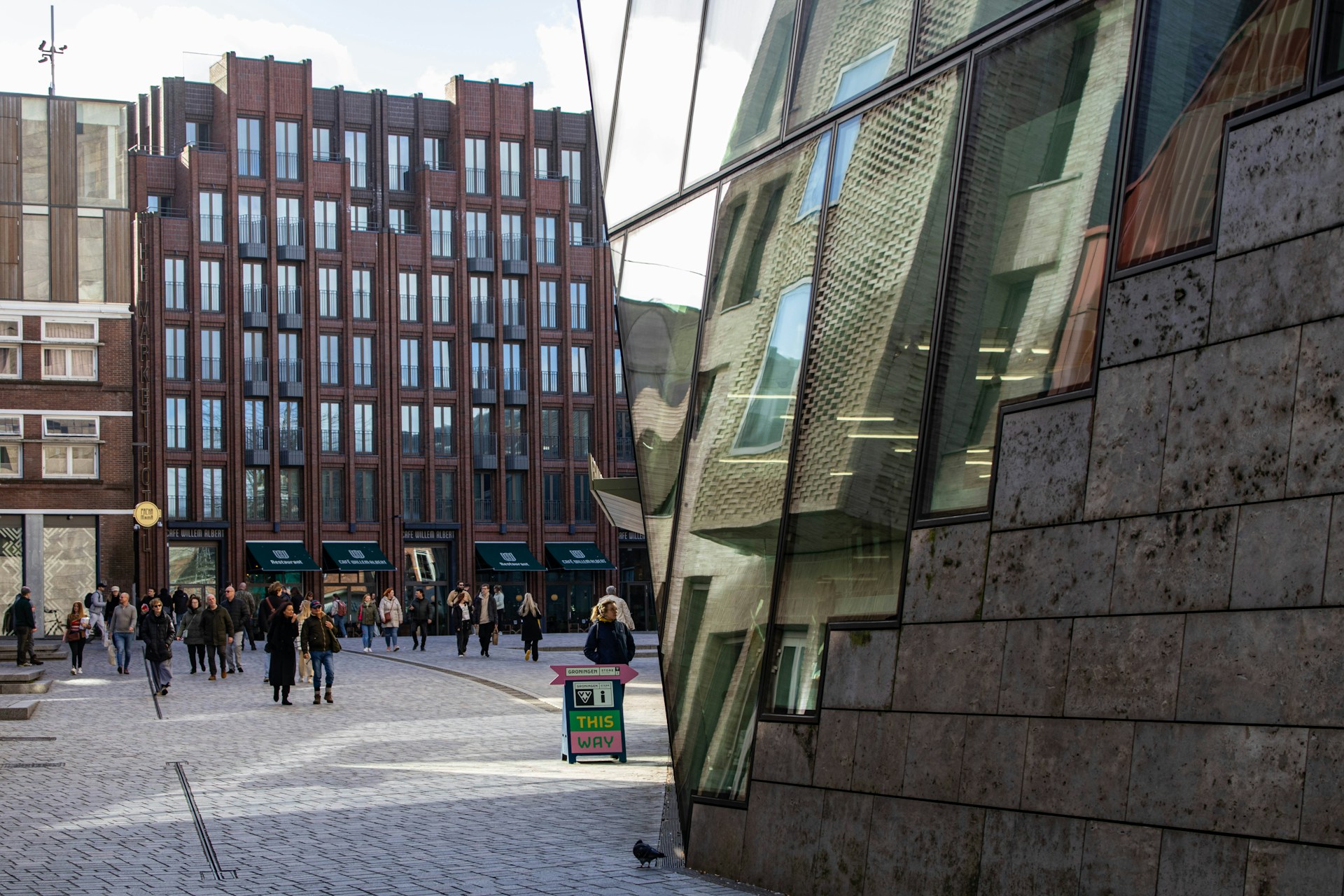 Een stadsplein met een aantal wandelende mensen en moderne gebouwen op de achtergrond, gedeeltelijk verborgen door digitale haperingen en vervormingen in het beeld.