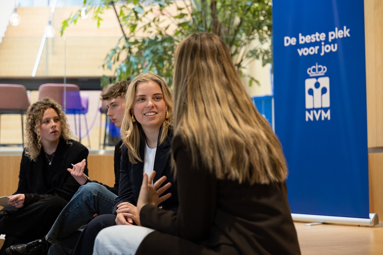Vier jongvolwassenen zitten binnen te praten bij een blauw bord met de tekst De beste plek voor jou en NVM, met op de achtergrond groen en een trap.