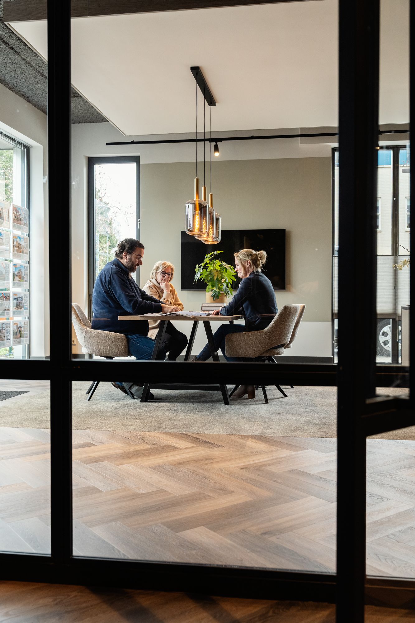 Drie mensen zitten rond een tafel in een modern kantoor en zijn in gesprek. Grote ramen laten natuurlijk licht binnen en een plant siert de kamer. De scène wordt bekeken door glazen wanden.